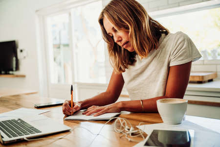 Caucasian student working from home writing on note pad with pen sitting with coffee and laptopの写真素材