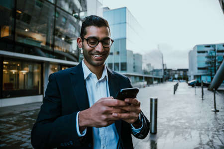 Mixed race businessman dressed smartly smiling while texting on smartphone walking on his way to...