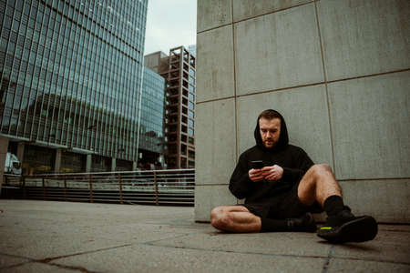 Caucasian male athlete sitting against wall typing on cellphone relaxing after long run