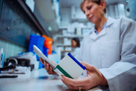 Elderly pharmacist reading label of prescription medication box standing behind counter in pharmacyの写真素材