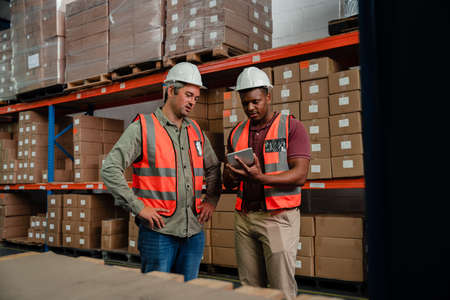 Male colleagues wearing overalls researching on digital tablet packaging processes standing in...