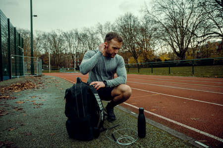 Caucasian male athlete putting earphones in crouching down next to backpack and waterbottleの写真素材