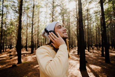 Handsome caucasian male wearing beanie and headphones smiling while listening to music standing in green forest.の写真素材