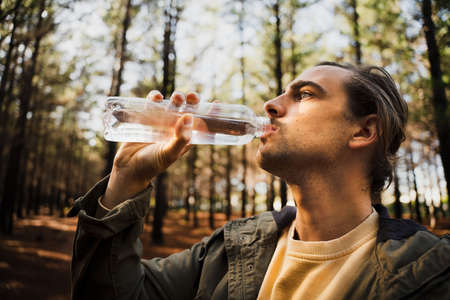 Young caucasian male drinking water from clear bottle in deep green forest.の写真素材
