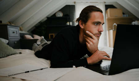 young caucasian male working from home typing on digital laptop relaxing in modern bedroomの写真素材