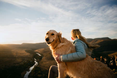 Blonde female sits on top of mountain with arm around golden puppy while looking into the distance at sunset.の写真素材