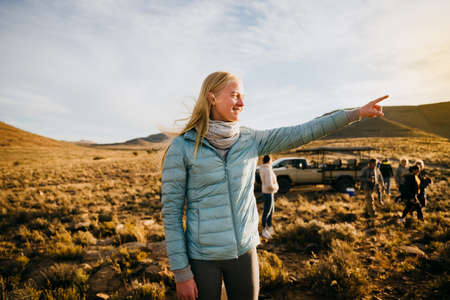 Beautiful caucasian lady point to the distance standing in green farmlands at sunset.