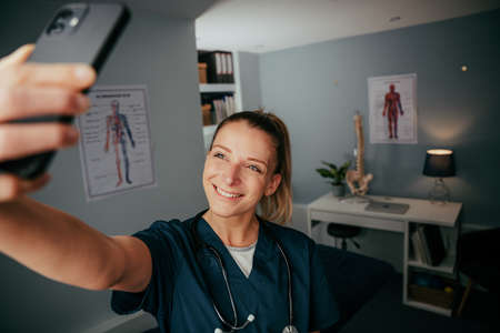 Caucasian female nurse standing in doctors office taking selfie on cellular device