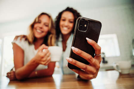 Beautiful caucasian and mixed race friends sitting in kitchen taking selfie on cellular device drinking hot coffeeの写真素材