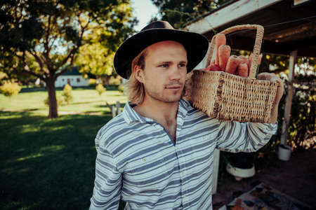 Handsome caucasian male focussed walking through fields in farm village holding basket of fresh vegetables and fruitの写真素材