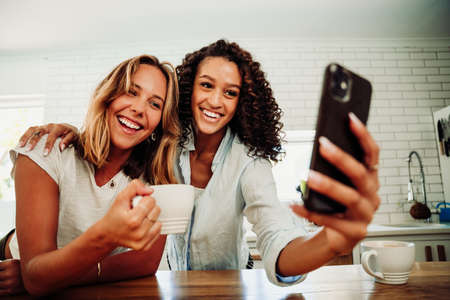 Mixed race female fridens sititng in kitchen taking selfies with cellular deviceの写真素材
