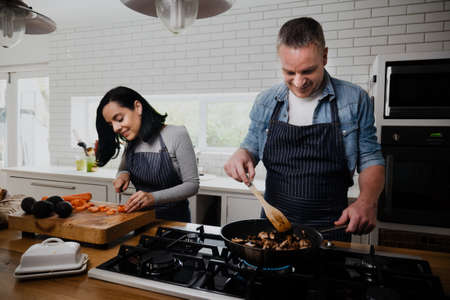 Happy couple preparing dinner while wife chops vegetables and husbands fries meat.の写真素材