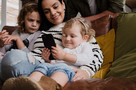 Mother and two daughters watching funny videos on smartphone sitting on sofa in modern lounge.の写真素材