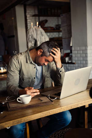 Stressed white male holding credit card ending laptop next to a coffee cup sitting in a rustic...