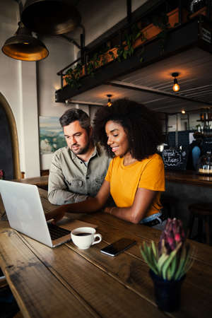 Smiling multi race couple browsing on laptop while drinking coffee in chic coffee shop.の写真素材