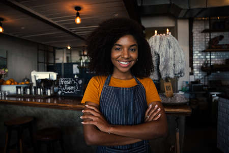 Beautiful smiling waitress wearing apron standing cross-armed in trendy cafeの写真素材