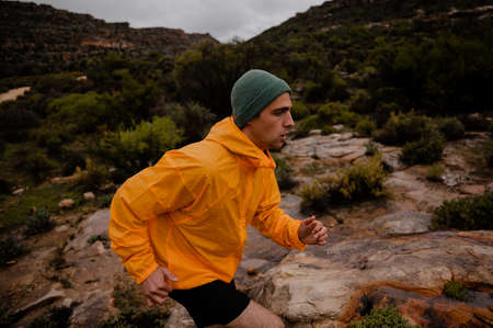 Tired fit teenage male sprinting up rocky mountain trail in wet weather with green bushes and steep hillsの写真素材