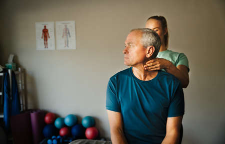 Male patient twisting his neck while female physiotherapist massages pinched nerves of neck standing in rehab room.の写真素材