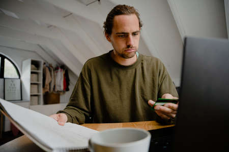 Male caucasian student working out monthly payments sitting at desk holding credit card and notepad...