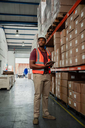 Concerned factory worker holding digital tablet organising parcels for delivery standing in...