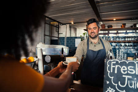 African American female collecting coffee from smiling handsome male waiter standing behind counter in stylish cafe.の写真素材