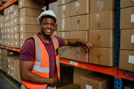 Male worker wearing hardhat leaning against parcels ready for delivery waiting in warehouseの写真素材