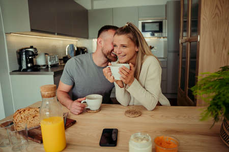 Caucasian happy couple sitting at kitchen table whispering in earの写真素材
