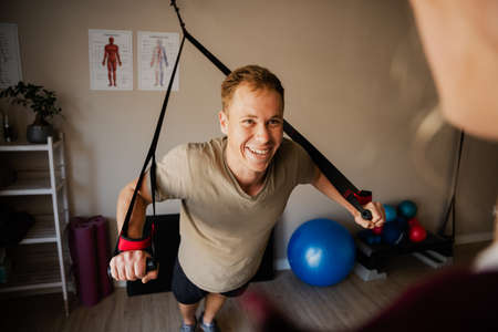 Smiling strong male patient leaning forward holding onto black rubber bands while female physiotherapist watches.の写真素材