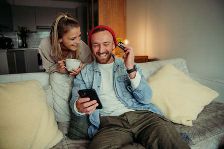 Caucasian couple sitting on couch drinking coffee making online payment using cellular device