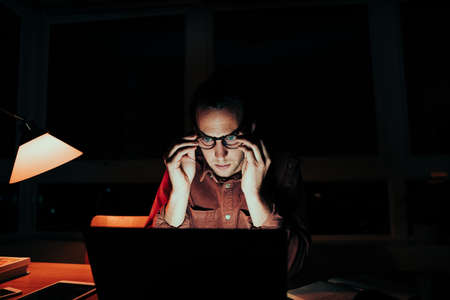 Stressed caucasian male freelancer adjusting spectacles working late at night on laptopの写真素材