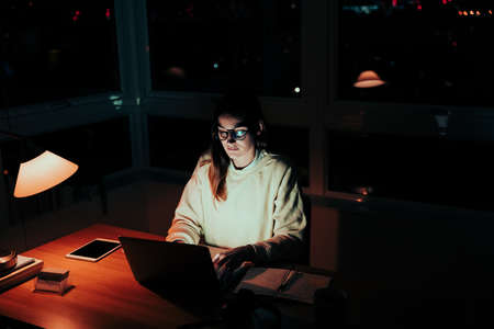 Caucasian female student working late in home office typing on laptop replying to emails from friends regarding homeworkの写真素材