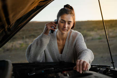 Afraid female fixing car after breaking down on highway, using smartphone to call for help.