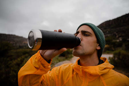 Tired young male trail runner drinking water from reusable bottle after running up steep mountain...