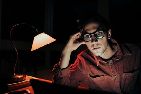 Caucasian male student working at laptop late at night tired leaning on hands meeting deadlinesの写真素材