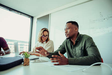 mixed race business man presenting ideas to clients in board meeting sitting besides female in board...