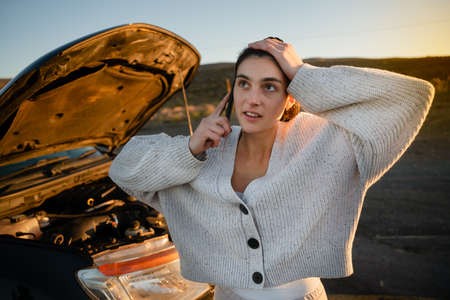 Stressed caucasian female using smartphone to call for help with broken down vehicle while standing alone on empty street.の写真素材