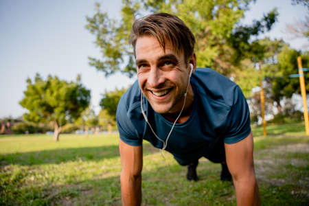Smiling handsome young male athlete planking exercise in sunny outdoor park ear phones playing musicの写真素材