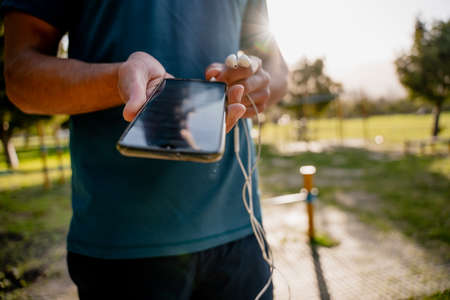 Young male athlete plugging ear phones into smartphone for music before exercising outdoors in sunny parkの写真素材