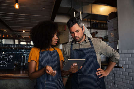 Curious colleagues wearing aprons, discussing finances while looking at tablet standing in coffee shop.の写真素材