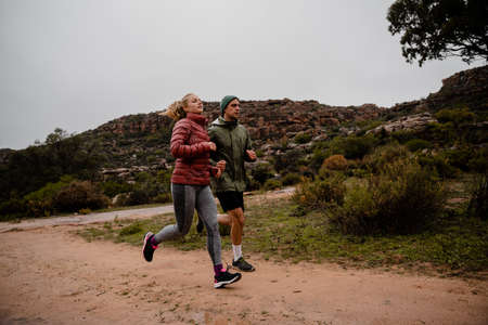 Strong fit young male and female athletes trail running on cold winters day on mountain path cloudy...