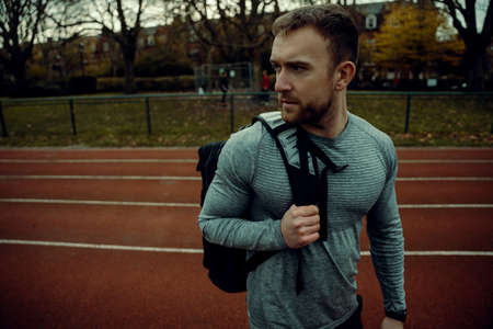 Caucasian male athlete walking on tracking field with backpack on hand looking awayの写真素材