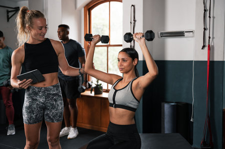 One woman lifts a dumbbell while the other uses a tablet at the gymの写真素材