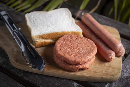 Fresh uncooked barbecue sausages, burgers and bread ready to be grilled, on wooden chopping boardの写真素材