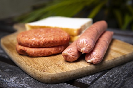 Fresh uncooked barbecue sausages, burgers and bread ready to be grilled, on wooden chopping boardの写真素材