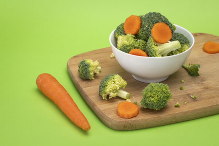 Broccoli and carrot mixed in white bowl sitting on wooden chopping boardの写真素材