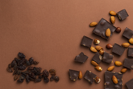 Dark chocolate pieces with roasted almonds and raisins scattered around on brown backgroundの写真素材