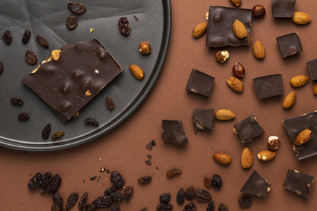 Dark chocolate pieces with roasted almonds and raisins scattered around on brown backgroundの写真素材