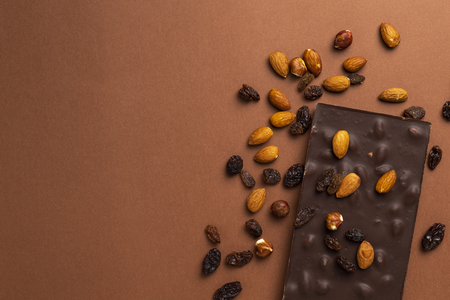 Dark chocolate bar with roasted almonds and raisins scattered around on brown backgroundの写真素材