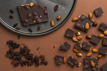 Dark chocolate pieces with roasted almonds and raisins scattered around on brown backgroundの写真素材