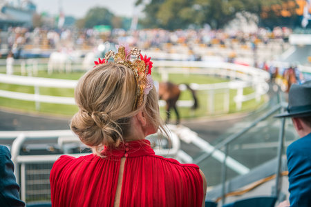 Woman in red dress wearing beautiful fascinator watching horses parading before the raceの写真素材
